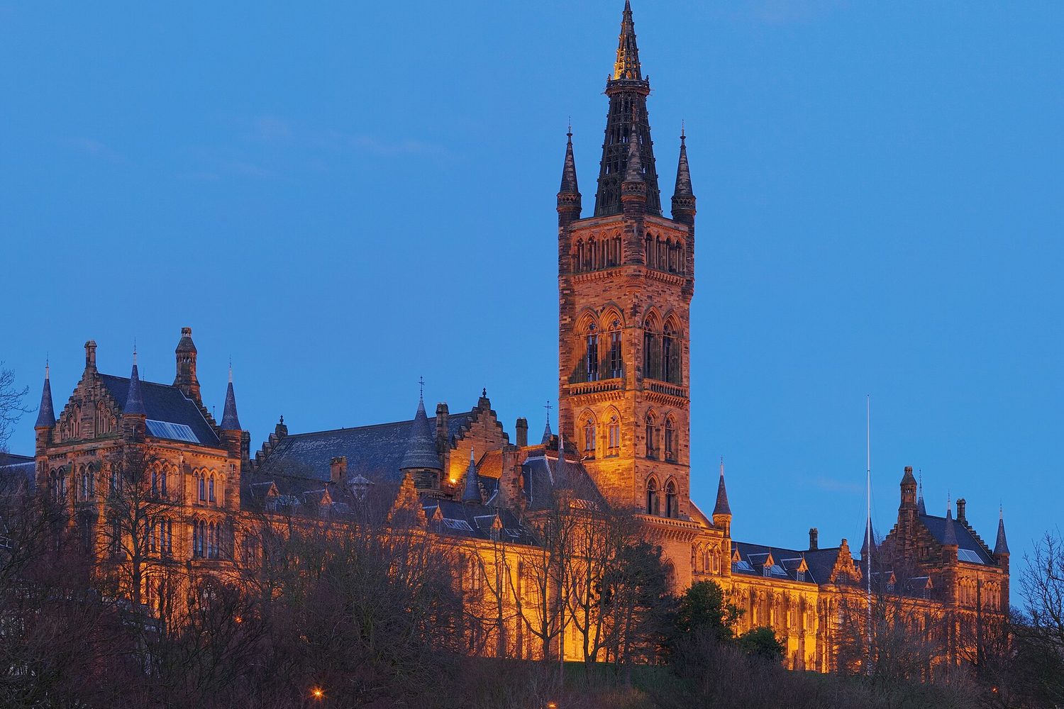 The Gilbert Scott Building at the University of Glasgow, floodlit at dusk, its tower and spire above the trees of Kelvingrove.